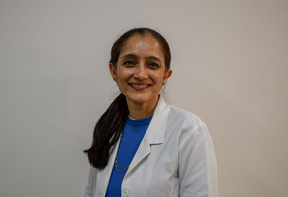The image shows a woman wearing a white coat standing against a plain background, smiling slightly and looking directly at the camera.