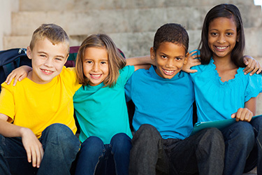 The image shows four children posing together with smiling faces  they are wearing blue shirts and are seated on a bench outdoors, possibly in front of a building.