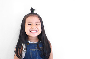 The image shows a young girl with dark hair smiling at the camera, wearing a denim jumper and standing against a plain background.
