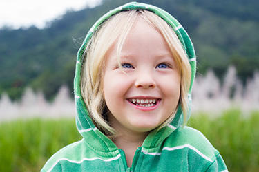 The image shows a young child smiling at the camera while wearing a green sweatshirt with a hood and a striped shirt underneath, against a blurred background of greenery.