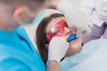 The image shows a dental professional performing a procedure on a patient s teeth while wearing protective eyewear and a surgical mask, with the patient seated in a dental chair and having their mouth opened by the dentist.