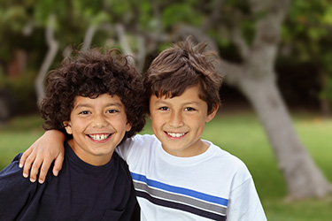The image features two young boys posing together with smiles, standing outdoors under a tree.