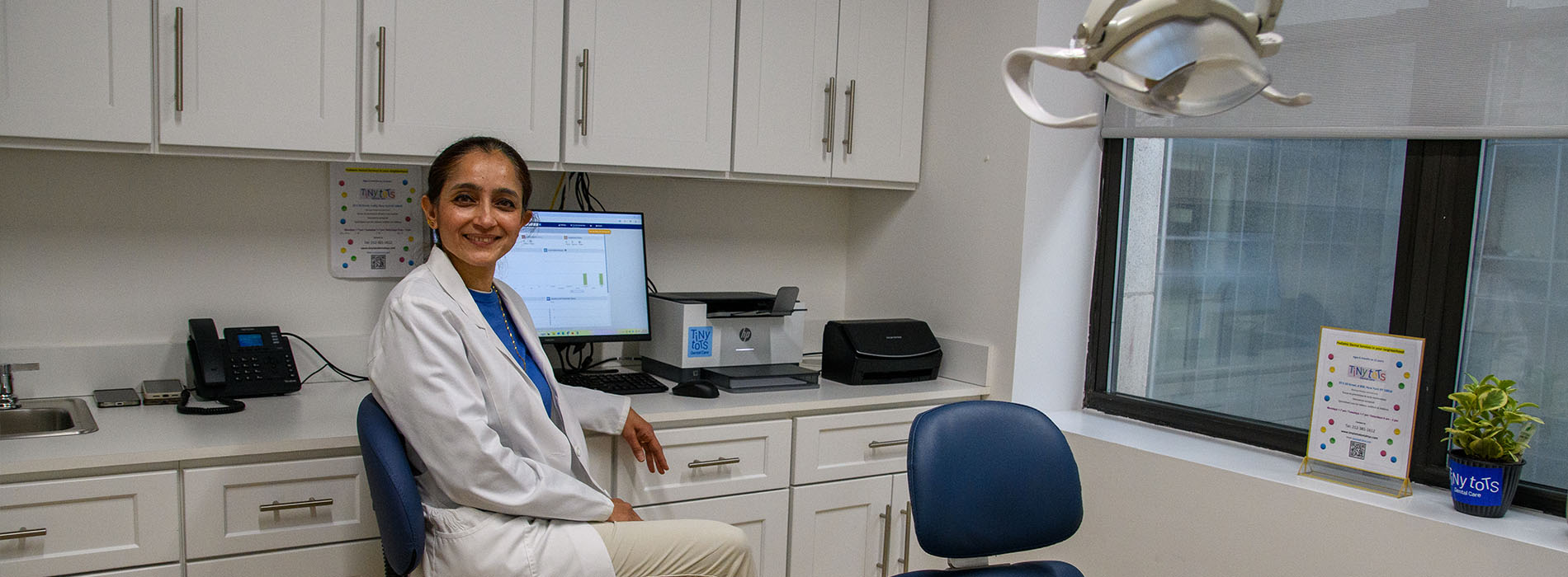 A woman sitting at a desk in a dental office, smiling towards the camera.