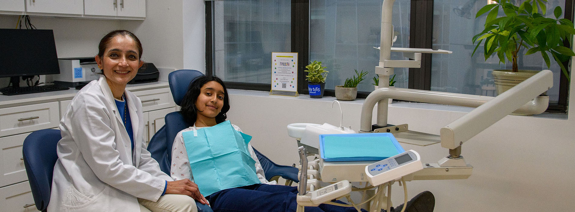 The image shows two individuals in a dental office setting, with one person seated on a dental chair and another standing beside them.