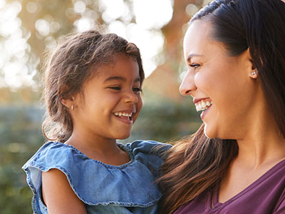 The image shows a woman and a young child smiling at each other, with the woman holding the child. They appear to be outdoors during daylight.